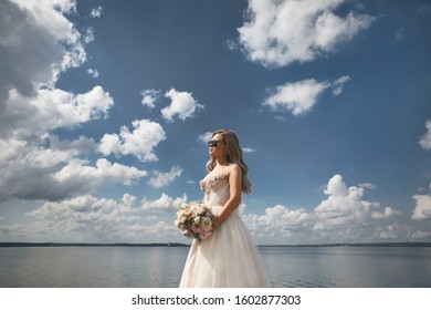 Fashionable Young Bride With Platinum Blond Hair In Wedding Dress Posing With Bouquet Of Fresh Flowers On The Sea Coast. Young Modish Woman With Wedding Hairstyle In Stylish Dress Posing Outdoors.