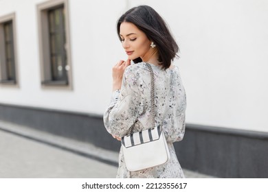 Fashionable young elegant pretty woman model with shorts hairstyle in stylish spring vintage dress with flowers print with fashion white bag walks on the city near a white wall - Shutterstock ID 2212355617