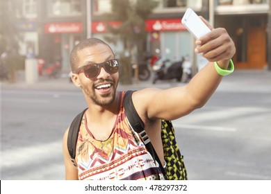 Fashionable Young Hipster Man Wearing Trendy Shades And Tank Top, Having Fun, Taking Self-portrait With Smart Phone Outdoors, Walking Down Street Alone On Sunny Summer Day. People And Technology