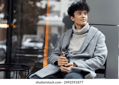 Fashionable young man in gray sweater and black pants sitting in chair with cup of coffee in front of glass window, looking off to side - Powered by Shutterstock