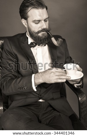 Fashionable young man with retro look sitting in an armchair, smoking pipe and holding a cup of coffee over gray background