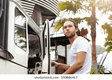 Fashionable young man with stylish beard wearing white t-shirt and baseball cap backwards looking away with confident and proud face expression while getting in his four-wheel drive vehicle - Powered by Shutterstock