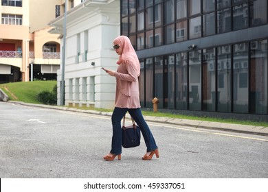 Fashionable Young Muslim Woman in Beautiful Hijab Busy texting with her Mobile Phone While Walking Across a City Street - Powered by Shutterstock