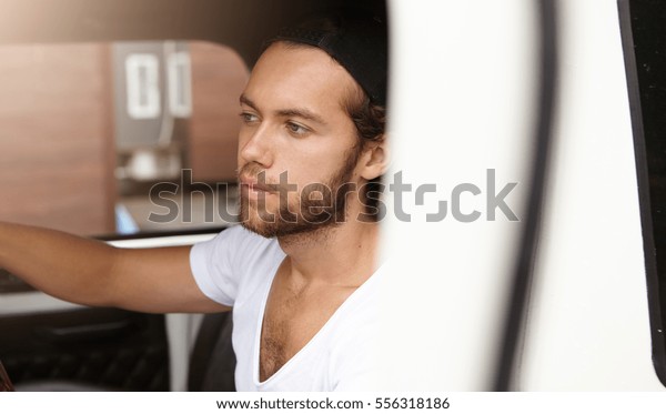 Fashionable young student wearing white t-shirt and\
snapback driving home after lectures at university, sitting inside\
leather cabin of his four-wheel drive car, having tired expression\
on his face