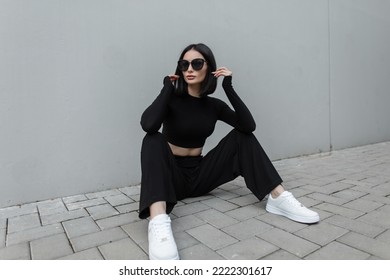 Fashionable young urban woman with black trendy sunglasses in fashion black stylish sports outfit with white sneakers sits on the street near a modern gray building - Powered by Shutterstock