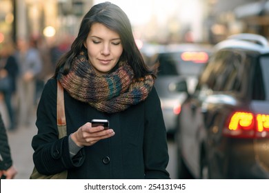 Fashionable Young Woman in Black Coat and Colorful Scarf Busy with her Mobile Phone While Walking a City Street