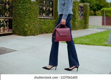 Fashionable Young Woman In Blue Flared Jeans, Grey Sweater, White Blouse And Burgundy Handbag .She Is Wearing Black High Heel Shoes. Street Style .