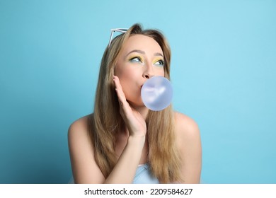 Fashionable Young Woman With Bright Makeup Blowing Bubblegum On Light Blue Background