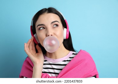 Fashionable Young Woman With Headphones Blowing Bubblegum On Light Blue Background