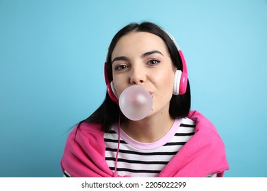 Fashionable Young Woman With Headphones Blowing Bubblegum On Light Blue Background