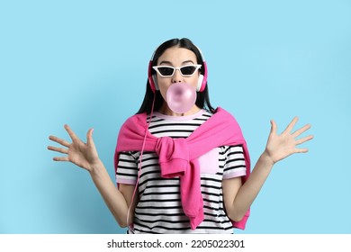 Fashionable Young Woman With Headphones Blowing Bubblegum On Light Blue Background