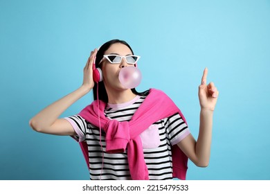 Fashionable Young Woman With Headphones Blowing Bubblegum On Light Blue Background