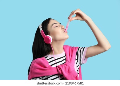 Fashionable Young Woman With Headphones Chewing Bubblegum On Light Blue Background