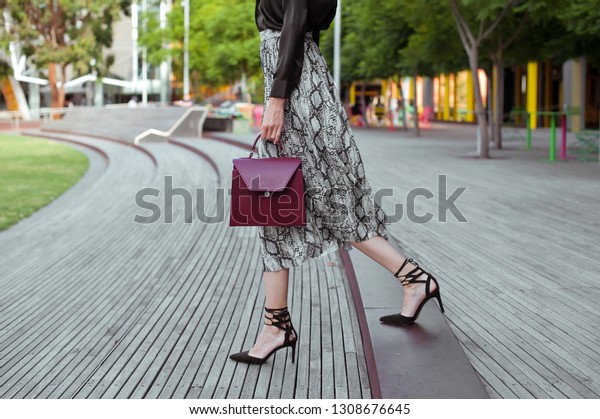 Fashionable young woman in high heel black shoes,\
black blouse, midi skirt with snake print and burgundy handbag in\
hand on the city street. Street\
style.