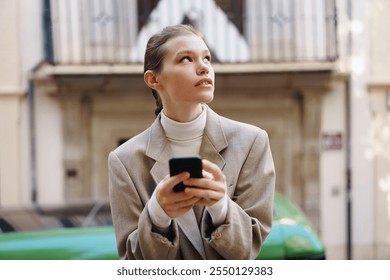 A fashionable young woman is holding a sleek smartphone, lost in thought as she gazes out into a vibrant urban environment, embodying a contemporary lifestyle that blends technology and modern living - Powered by Shutterstock