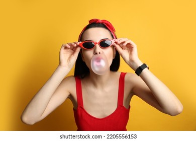 Fashionable Young Woman In Pin Up Outfit Blowing Bubblegum On Yellow Background