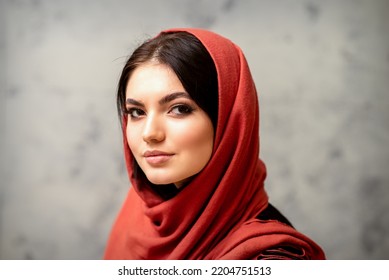 The Fashionable Young Woman. Portrait Of The Beautiful Female Model With Long Hair And Makeup In A Red Scarf. Beauty Young Caucasian Woman On The Background Of A Gray Wall