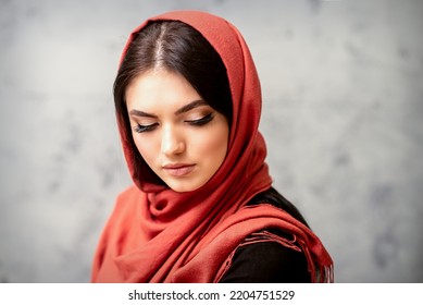 The Fashionable Young Woman. Portrait Of The Beautiful Female Model With Long Hair And Makeup With Eyelash Extensions In A Red Scarf. Beauty Young Caucasian Woman On The Background Of A Gray Wall