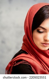 The Fashionable Young Woman. Portrait Of The Beautiful Female Model With Long Hair And Makeup With Eyelash Extensions In A Red Scarf. Beauty Young Caucasian Woman On The Background Of A Gray Wall