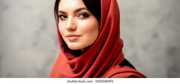 The Fashionable Young Woman. Portrait Of The Beautiful Female Model With Long Hair And Makeup In A Red Scarf. Beauty Young Caucasian Woman On The Background Of A Gray Wall