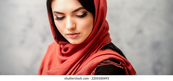 The Fashionable Young Woman. Portrait Of The Beautiful Female Model With Long Hair And Makeup With Eyelash Extensions In A Red Scarf. Beauty Young Caucasian Woman On The Background Of A Gray Wall