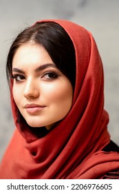 The Fashionable Young Woman. Portrait Of The Beautiful Female Model With Long Hair And Makeup In A Red Scarf. Beauty Young Caucasian Woman On The Background Of A Gray Wall