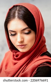 The Fashionable Young Woman. Portrait Of The Beautiful Female Model With Long Hair And Makeup With Eyelash Extensions In A Red Scarf. Beauty Young Caucasian Woman On The Background Of A Gray Wall