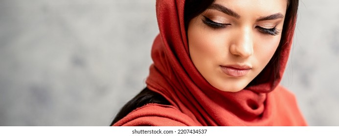 The Fashionable Young Woman. Portrait Of The Beautiful Female Model With Long Hair And Makeup With Eyelash Extensions In A Red Scarf. Beauty Young Caucasian Woman On The Background Of A Gray Wall