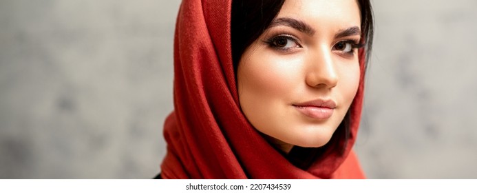 The Fashionable Young Woman. Portrait Of The Beautiful Female Model With Long Hair And Makeup In A Red Scarf. Beauty Young Caucasian Woman On The Background Of A Gray Wall