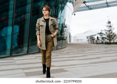 Fashionable Young Woman With Short Hairstyle Wearing Green Suit And Stylish Glasses Walking In Outdoor Business District And Looking Away.