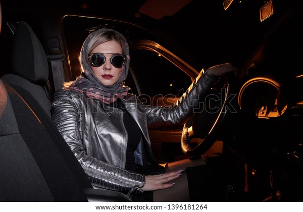 Fashionable young woman in a\
silver leather jacket in a car in the black night and colour light\
behing her