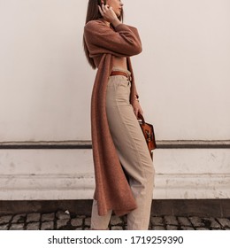 Fashionable Young Woman In Stylish Beige Trousers In Long Vintage Coat With Leather Brown Trendy Handbag Posing Near White Wall In City. Closeup Female Body In Elegant Clothes. Spring Womens Fashion.