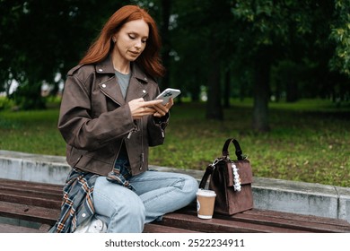 Fashionable young woman with vibrant red hair relaxing on wooden bench in beautiful urban park, pausing from freelancing to use smartphone and savor cup of coffee. Stylish redhead woman relax alone. - Powered by Shutterstock