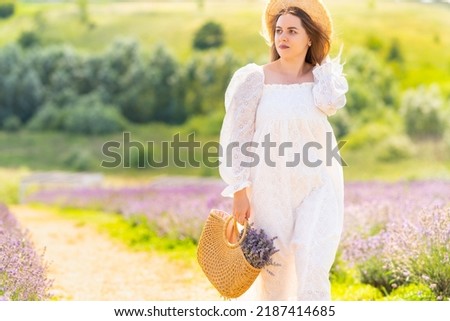 Fashionable young woman walking through rural lavender fields in a white summer frock and straw hat