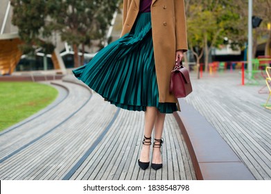 Fashionable young woman wearing green pleated midi skirt, sweater, high heel shoes, beige wool coat and holding burgundy handbag in hand on the city street. Street style.