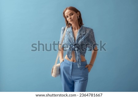 Fashionable young woman wearing  knotted chiffon blouse, trousers, carrying trendy white leather padded cassette shoulder bag, posing on blue background. Studio portrait. Copy, empty space for text