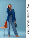 Fashionable young woman wearing trendy orange sunglasses, blue linen shirt, trousers, block heel shoes, holding bag, posing on blue background. Full-lenght studio portrait. Copy, empty space for text
