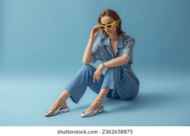 Fashionable young woman wearing trendy yellow sunglasses, blue chiffon blouse, trousers, silver ballet flats, posing on blue background. Full-length studio portrait. Copy, empty space for text