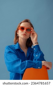 Fashionable young woman wearing trendy orange sunglasses, linen shirt, posing on blue background. Studio portrait. Copy, empty space for text  - Shutterstock ID 2364781657