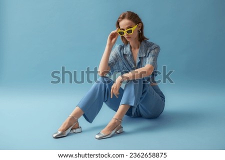 Fashionable young woman wearing trendy yellow sunglasses, blue chiffon blouse, trousers, silver ballet flats, posing on blue background. Full-length studio portrait. Copy, empty space for text