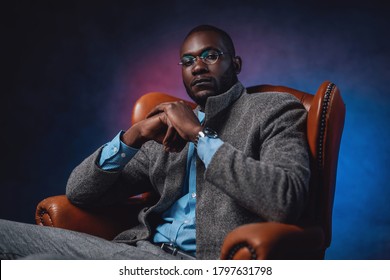 Fashoinable stylish blackman are sitting on a chair with multicolored background behind. Business lifestyle. Expensive custom suit.