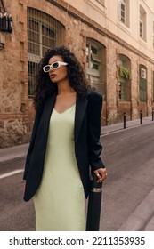 Fasionable young african girl in stylish sunglasses looking into distance stands on europe street. Brunette with curly hair wears coctail dress and black jacket. Concept fasion, female beauty.