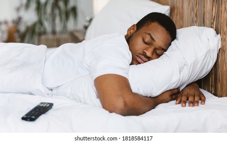 Fast Asleep Black Man Sleeping Embracing Pillow Resting In Bed At Home, Lying Near Remote Control Tired After Bingewatching TV Shows At Night. Rest, Napping And Recreation Concept.