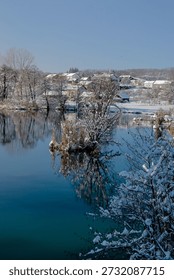 A fast, cold river flows through a winter landscape on a bright, sunny day. Snow blankets the ground, covering nearby pine trees, small houses, and quiet roads that wind gently through the scene. The dark, moving water reflects the snow-covered pine trees along the riverbanks, creating shimmering reflections that add depth and visual balance. Sunlight highlights icy edges and rippling currents, enhancing the contrast between motion and stillness.

In the distance, simple houses rest peacefully among the trees, their roofs layered with snow, suggesting warmth and shelter during the cold season. Snow-covered roads lead through the landscape, guiding the eye and adding a clear sense of place. The air feels crisp and fresh, reinforcing the clean, natural atmosphere.

This winter scene combines natural energy with subtle human presence, making it ideal for themes such as seasonal travel, rural life, winter lifestyle, resilience, and European countryside beauty.
