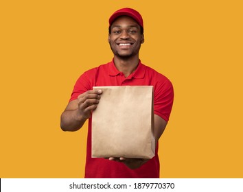 Fast Delivery. Front View Of Smiling African American Courier Guy In Red Cap And Uniform Holding And Giving Craft Paper Bag From Restaurant With Products To Camera, Yellow Orange Studio Background