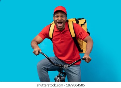 Fast Delivery Service. Overjoyed african american courier guy in red cap and uniform riding bicycle and wearing thermo backpack bag isolated on blue studio background. Shipment service during lockdown