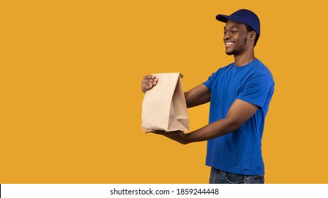 Fast Delivery. Side View Of Smiling African American Delivery Guy In Blue Cap Holding And Giving Craft Paper Bag From Restaurant With Products To The Side With Copy Space, Orange Background