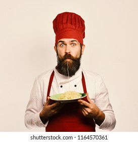 Fast food concept. Chef eats italian or asian noodles. Man with beard and noodle in mouth holds tasty dish on white background. Cook with shocked face in burgundy uniform holds fork and plate.