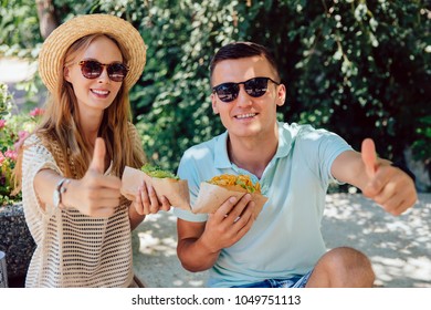 Fast Food Concept. Smiling Attractive Man And Woman Holding Hot Dog, Showing A Thumb Up, Looking At Camera. Dressed In Summer Clothes, In Sunglasses.