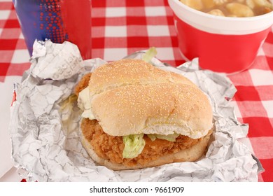 Fast Food Crispy Chicken Burger Still In Wrapper With With Drink And Poutine On A Checkered Red Background